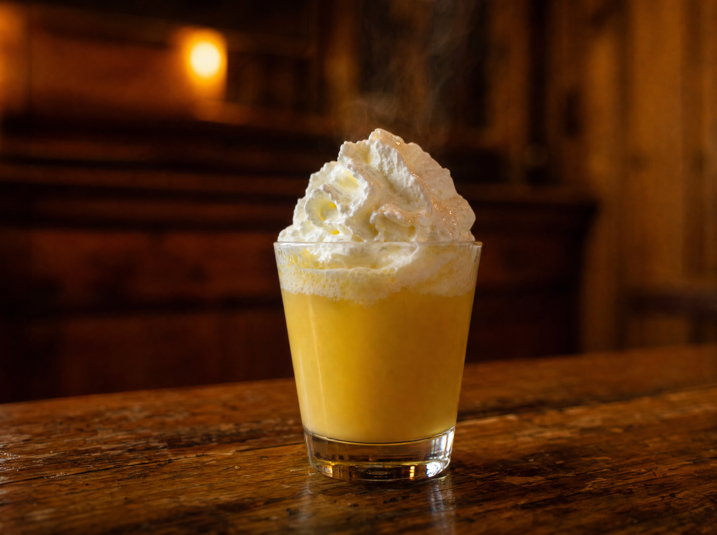 A heatproof glass of warm golden bombardino topped with whipped cream and dusted with cocoa.