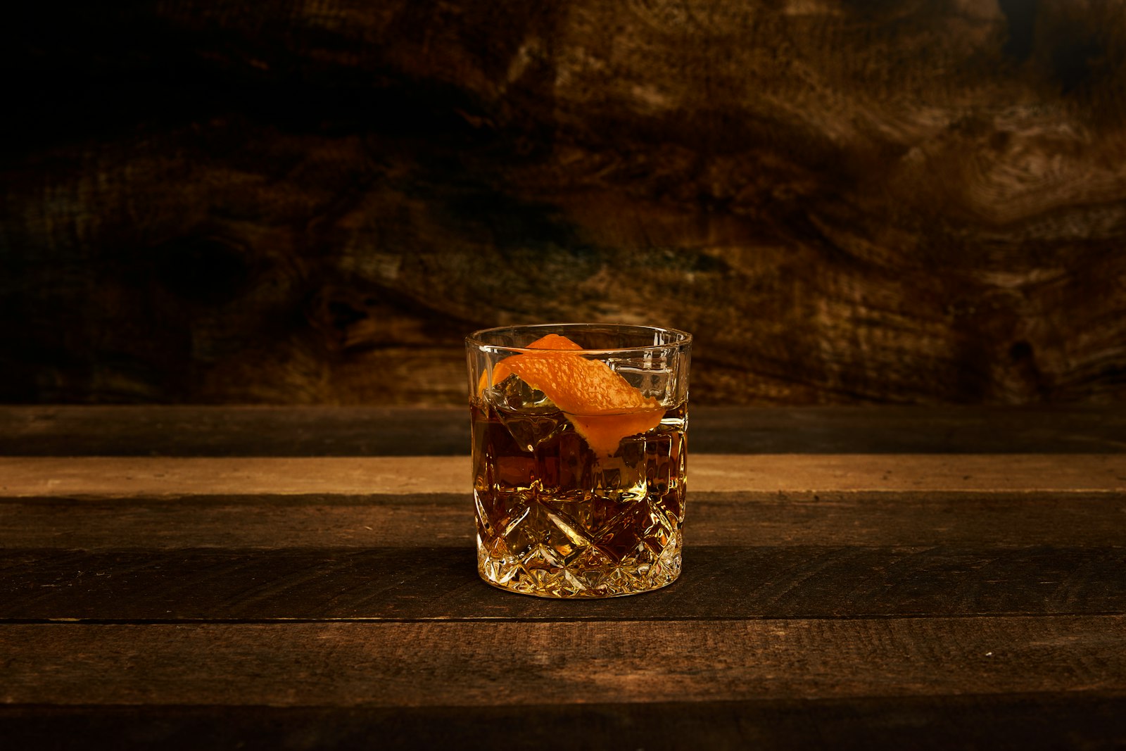 Chai spiced old fashioned in a rocks glass with a single large ice cube, orange twist and cinnamon stick, warm amber colour.