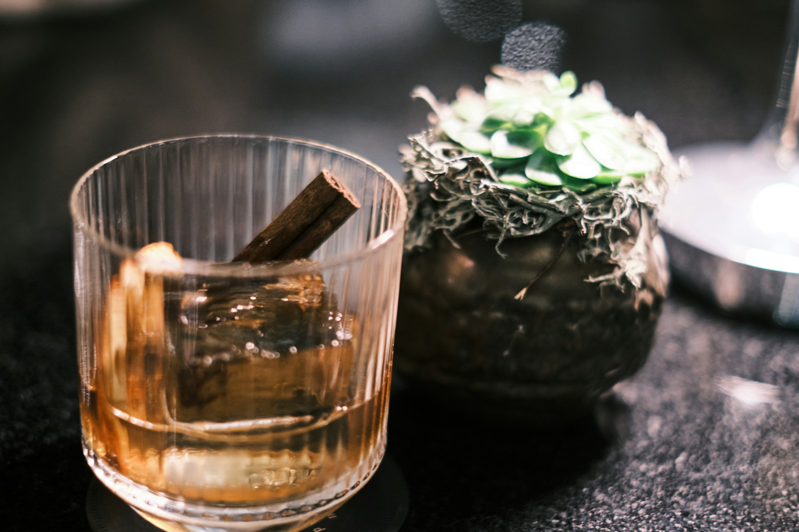 Cinnamon old fashioned in a heavy rocks glass with a large clear ice cube, orange peel and cinnamon stick garnish.
